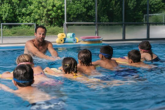 Schwimmkurs mit Lehrer und Kindergruppe &ndash; Ein Schwimmlehrer &uuml;bt mit mehreren Kindern im Freibad den sicheren Umgang mit Schwimmbrettern, lebendige Szene mit Bewegung und Wasserspritzern.