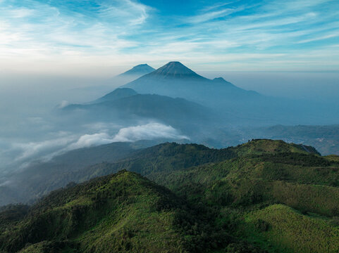 Aerial view of Gunung Prau surrounded by lush green hills, rolling clouds, and mist under a clear blue sky in Bawang, Central Java, Indonesia.