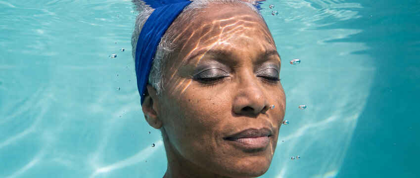 Underwater woman floating peacefully in pool features a serene senior swimmer with closed eyes, surrounded by clear blue water, sunlight caustics, and air bubbles for wellness content