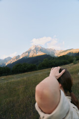 Obraz premium A young woman in a white shirt stands in a mountain meadow with her hand behind her head, gazing toward distant peaks.