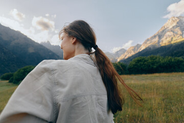 Obraz premium A woman with long hair tied in a ponytail stands outdoors in a grassy field surrounded by mountains under a partly cloudy sky, enjoying a peaceful lifestyle moment.
