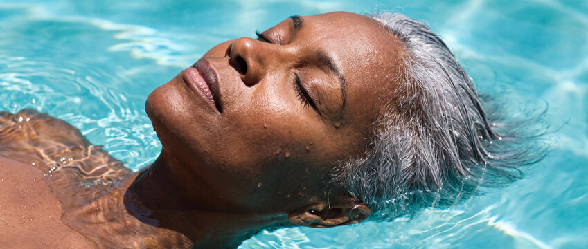 Senior woman floating in clear swimming pool water with eyes closed and silver hair, bathed in bright sunlight with turquoise reflections for senior wellness and retirement concept