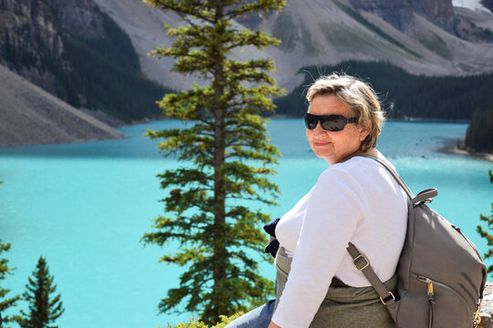 Side view of senior woman with backpack and sunglasses posing in front of turquoise mountain lake with evergreen tree and rocky mountain background