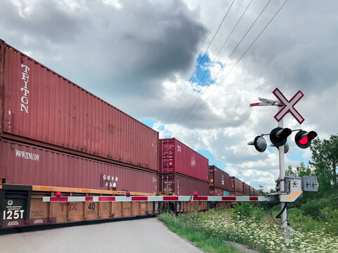 Long freight train with red TRITON and ZIM shipping containers passing railroad crossing with red signal light and crossing gate under cloudy sky, Ontario Canada 2020