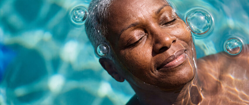 Senior woman relaxing in swimming pool with closed eyes and water bubbles around her face, captured in bright sunlight for wellness, spa, mental health, and active retirement concepts