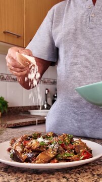 Unrecognizable older man adding grated parmesan cheese to a traditional Italian eggplant parmigiana dish, preparing a healthy vegetarian meal at home in his kitchen