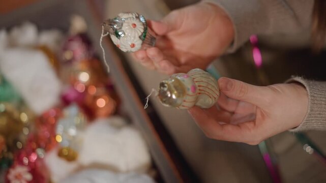 Hands holding vintage glass ornament gently, caucasian woman inspects fragile bauble in cozy living room box with tissue paper, warm bokeh lights, careful fingers arranging sparkle and tinsel, tender