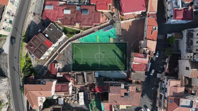 Aerial view of a green soccer field surrounded by dense residential buildings with terracotta rooftops in the coastal town of Cetara, Amalfi Coast, Campania, Italy.