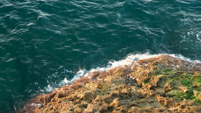 Aerial top down view of turquoise sea waves splashing against rocky coastline with orange rocks and green algae on a sunny day