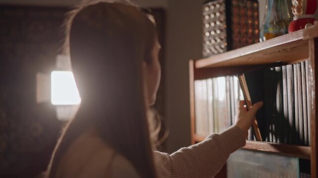 White woman examining bookshelf carefully. Young lady with white skin exploring vintage book spines warmly. Teenage female wearing sweater selecting wellloved books in cozy cinematic setting
