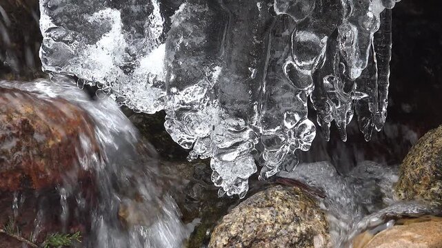 Icicle ice forms above flowing stream water between stones in cold winter nature scene.