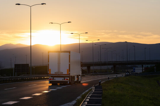 White cargo truck driving on a highway at sunset, rear view, with road signs, street lights and mountains silhouetted against the golden evening sky