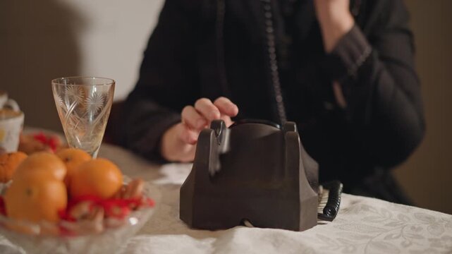 Person talking on vintage telephone at dinner table, receiver held to ear with cord trailing across lace tablecloth, fruit bowl and crystal glass in foreground, soft shadows and close framing convey