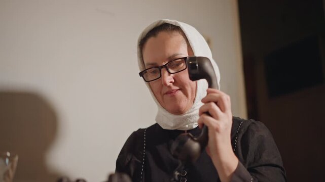 White woman in headscarf dialing telephone, intimate domestic closeup showing glasses and vintage handset, slow deliberate motions, scene suggests grandmother, neighbor, prayerful caller, memory