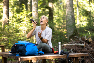 Fototapeta premium Happy woman sitting on a wooden bench, laughing while talking on her phone during a hiking trip in the green forest