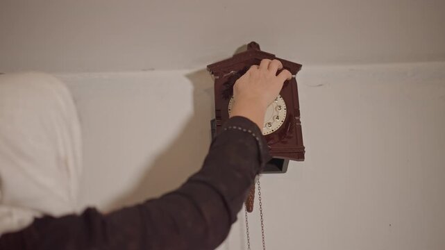 White matriarch reinstalls family heirloom clock on living room wall, careful hands align pendulum and chains, soft home lighting and vintage wood case create warm nostalgic atmosphere