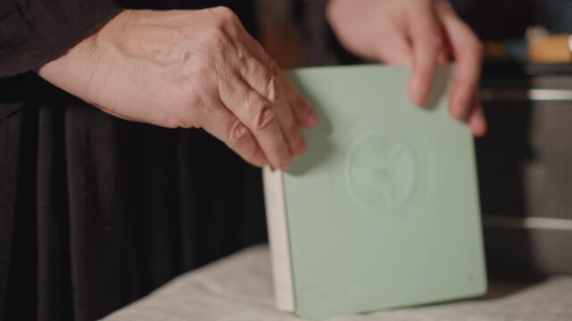 Closeup hands holding mint archive book, gentle fingers trace embossed emblem on cover, researcher catalogs object at table, quiet library atmosphere filled with preservation and memory.