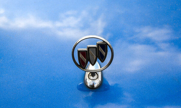 Close-up of a classic chrome Buick tri shield hood ornament mounted on a blue car bonnet, with a subtle reflection of the sky and light clouds