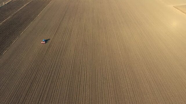 Aerial view of blue tractor sowing seeds across wide flat brown agricultural plain landscape.