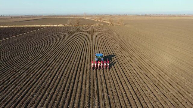 Aerial view of blue tractor planting fertilizer and seeds across wide brown field farmland.
