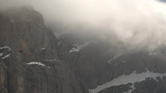 Sharp cliff peak rises from snowy rocky high mountain wall surrounded by dense fog layers.