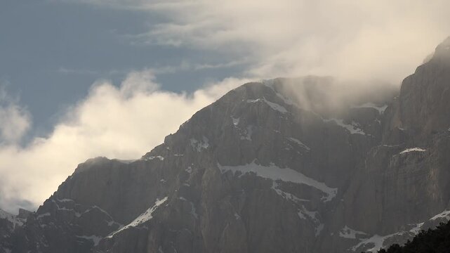 Sharp cliff peak rises from snowy rocky high mountain wall surrounded by dense fog layers.