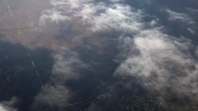 Aerial airplane window scene shows patchy clouds over farmland, forest, and distant city landforms.