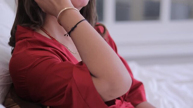 Close up of a woman wearing a luxurious red silk robe and a delicate necklace, posing in a soft, natural light bedroom setting.