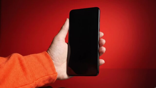 Closeup of a hand holding a modern smartphone with a blank black screen against a solid red background