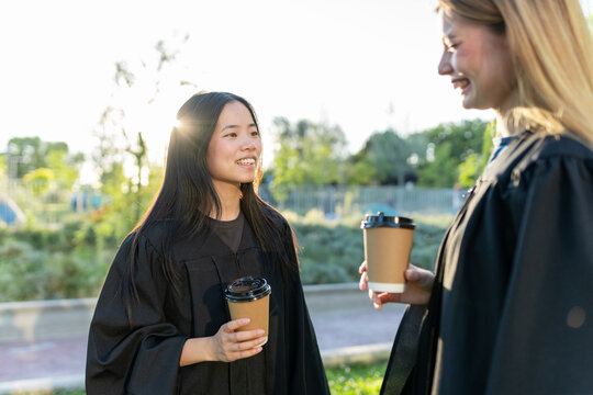 Graduating students wearing academic gowns, smiling and talking outdoors while holding takeaway coffee cups