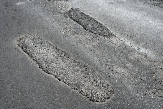 Close-up of a damaged asphalt road surface featuring rough patched potholes, cracks, and uneven pavement in bright daylight.