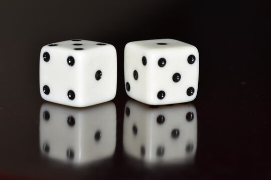 A pair of white, six-sided dice resting on a glossy black surface with a clear reflection, displaying a total sum of seven (a four on the left die and a three on the right die)