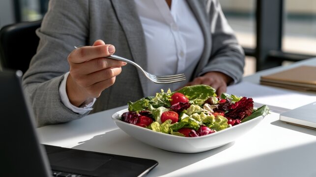 Business professional holds fork over fresh salad in bowl at desk. Laptop and notebook sit nearby during lunch break in office