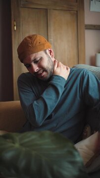 Young Latino man sitting on his sofa with significant neck pain caused by poor posture, visibly uncomfortable while holding or rubbing his neck.
