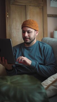 Young Latino man sitting on his sofa using a laptop, representing technology, remote work, productivity, digital communication, and everyday life at home.
