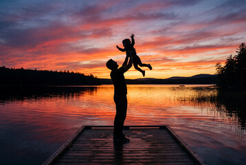 Fototapeta premium Silhouette of a father lifting his child in the air standing on a wooden lake dock at sunset