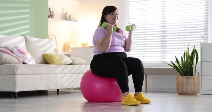 Overweight woman exercising with dumbbells on fitness ball indoors. Sports equipment