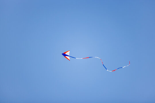 A small red, white and blue kite flies with a long striped tail against a clear light blue sky.