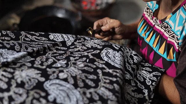 Elderly Indonesian artisan hand using a traditional canting tool to apply hot liquid wax on fabric, the process of making batik