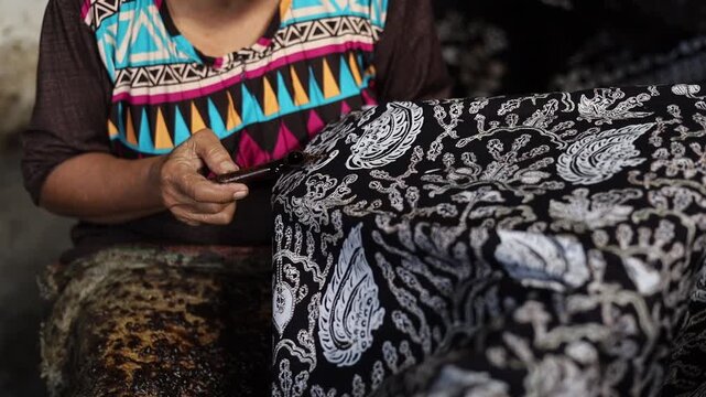Close up of Elderly Indonesian artisan hand using a traditional canting tool to apply hot liquid wax on fabric. Authentic process of making handmade batik textile.