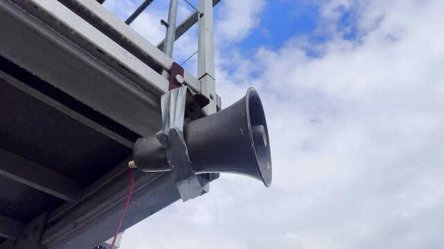 Close-up of a megaphone speaker mounted under a metal platform, with taped bracket, red wiring, scaffold rails and cloudy blue sky background.