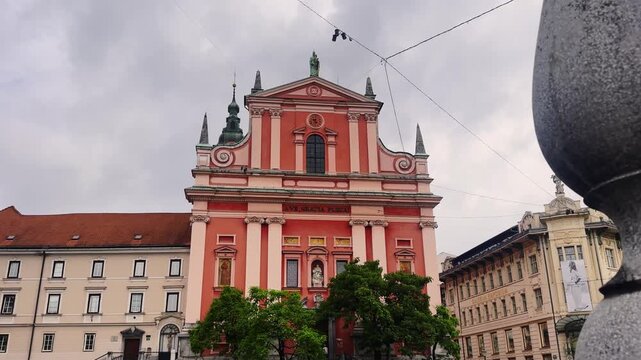 Low-angle view of the Franciscan Church of the Annunciation on Pre&scaron;eren Square, with its pink Baroque facade, historic buildings and a stone monument base in Ljubljana.
