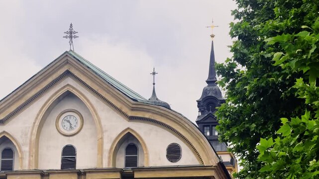 Close telephoto view of the Ursuline Church of the Holy Trinity in Ljubljana, showing its curved clock gable and dark spires framed by green leaves under an overcast sky.