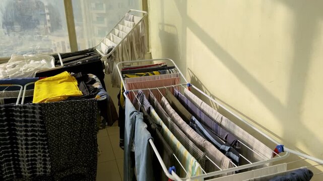 Various pieces of clothing hanging on folding drying racks inside a sunlit home. The organized laundry dries naturally near a window during the daytime, representing daily domestic chores and housekee