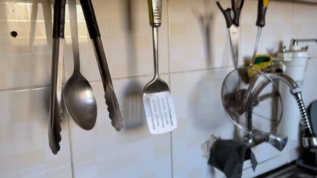 Close-up of metallic kitchen utensils including tongs, spoon, and spatula hanging on a beige tiled wall. Everyday cooking tools stored in a domestic home kitchen.