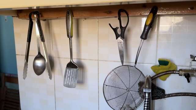 Metal cooking utensils hang from hooks above a kitchen sink and tiled backsplash, showing organized food-preparation tools in a naturally lit home interior.