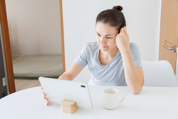 Woman using a tablet while sitting at a table with a cup of coffee at home. Digital technology, online communication, remote work and everyday lifestyle. © soya