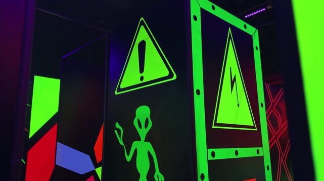 Fluorescent alien graphics and electrical warning signs glow under blacklight inside a dark indoor play area, creating a colorful sci-fi gaming atmosphere.