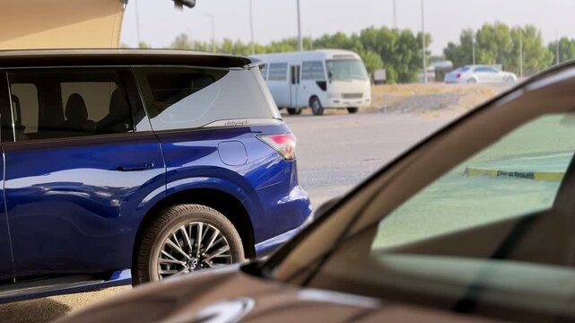 A dark blue SUV is parked in an outdoor parking lot during daytime. A blurred car sits in the foreground, with a white minibus, a distant sedan, and green trees visible in the sunny background.