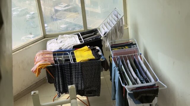 High-angle view of freshly washed clothes hanging on folding drying racks inside a bright stairwell landing. Strong daylight streams through a large window overlooking parked cars and buildings.